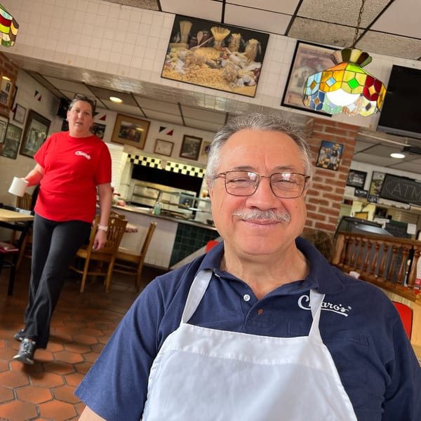 Smiling man in a Chiaro's shirt and apron, with a woman in a red shirt in the background.
