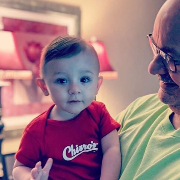 A baby in a red Chiaro's Pizzeria shirt sits on an older man's lap.