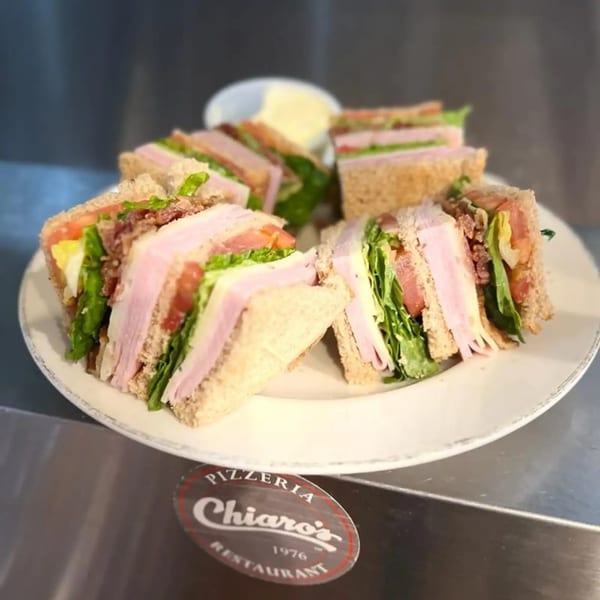 A white plate holds several club sandwich halves with ham, cheese, lettuce, tomato, and bacon.